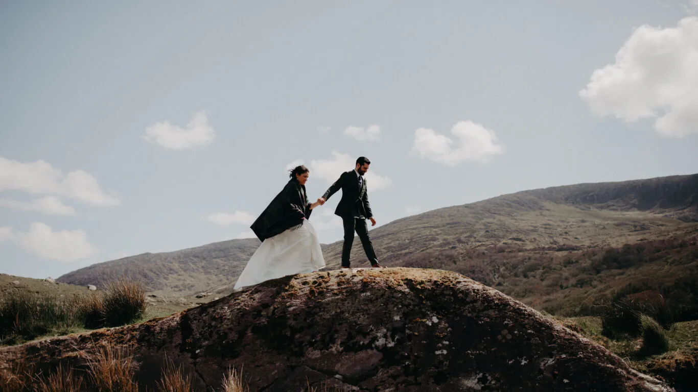 A bride and groom in formal attire walk hand-in-hand on a rocky hill with mountains and a cloudy sky in the background.