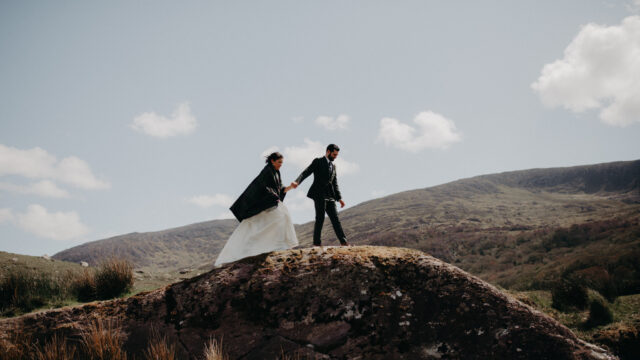 A bride and groom in formal attire walk hand-in-hand on a rocky hill with mountains and a cloudy sky in the background.