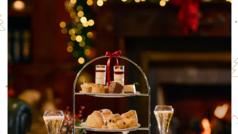 A festive afternoon tea setup with a three-tiered tray of pastries and sandwiches, glasses of sparkling wine, and holiday decorations in the background.