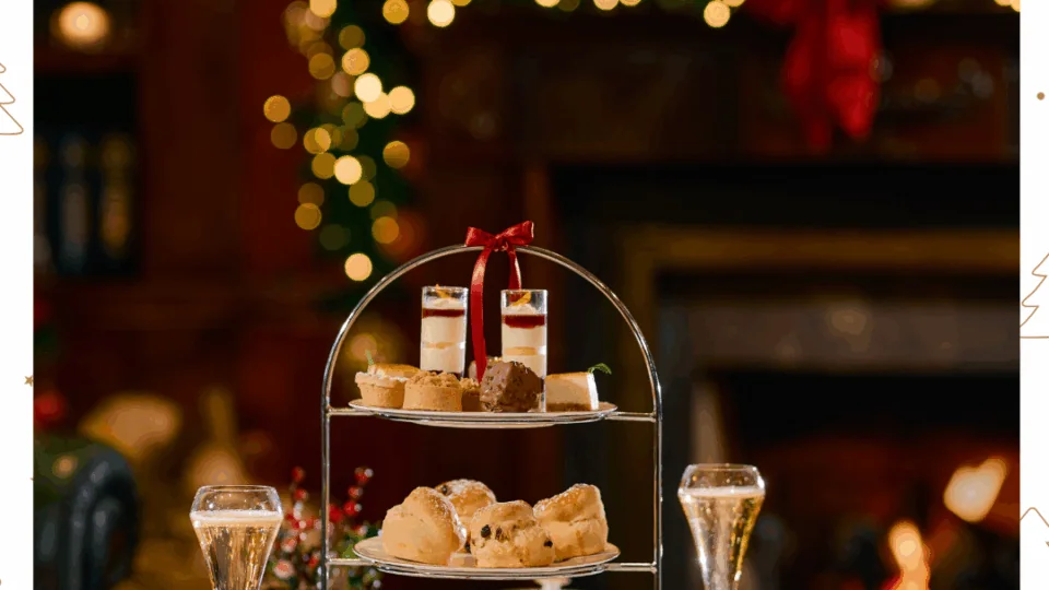 A festive afternoon tea setup with a three-tiered tray of pastries and sandwiches, glasses of sparkling wine, and holiday decorations in the background.