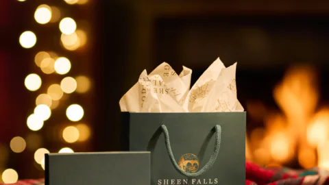 A Sheen Falls Lodge gift bag and box with tissue paper, surrounded by holly, are displayed in front of festive lights and a fireplace.