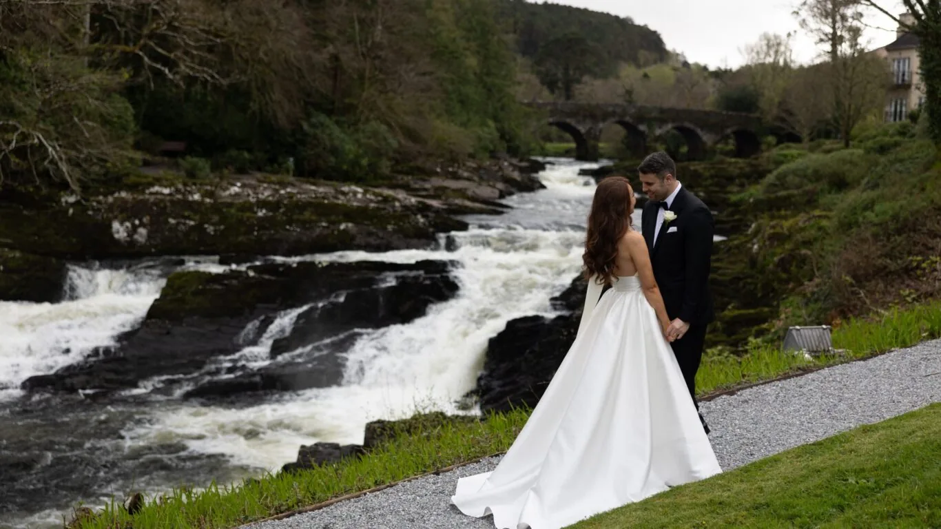 A bride and groom in formal attire stand hand in hand near a river with rapids, surrounded by greenery and a stone bridge in the background.