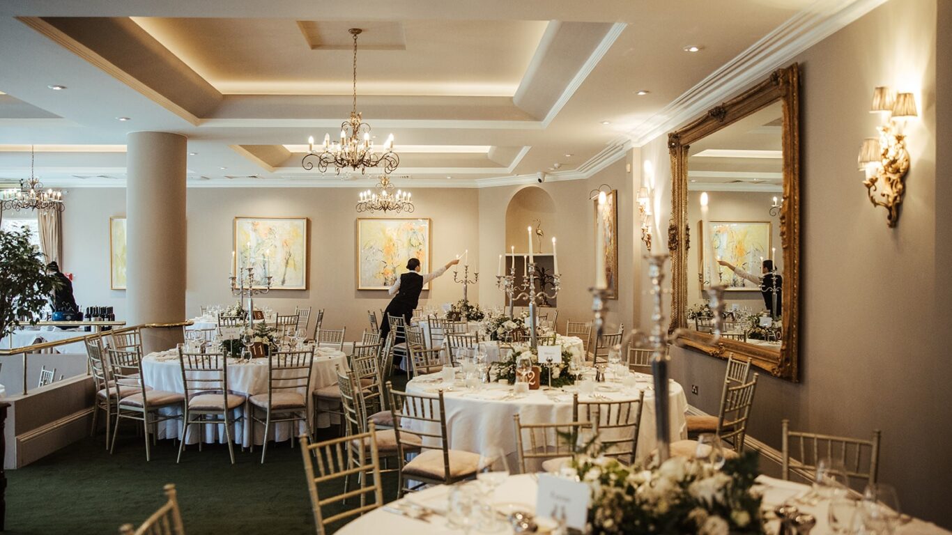 A formal banquet hall with round tables set for an event, decorated with white flowers and candles. A person arranges items on one table. Chandeliers and art adorn the space.