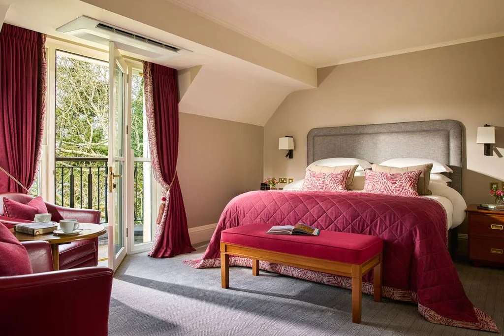 Elegant hotel room with king-size bed, red quilt, cosy armchairs, balcony view, and abundant natural light.