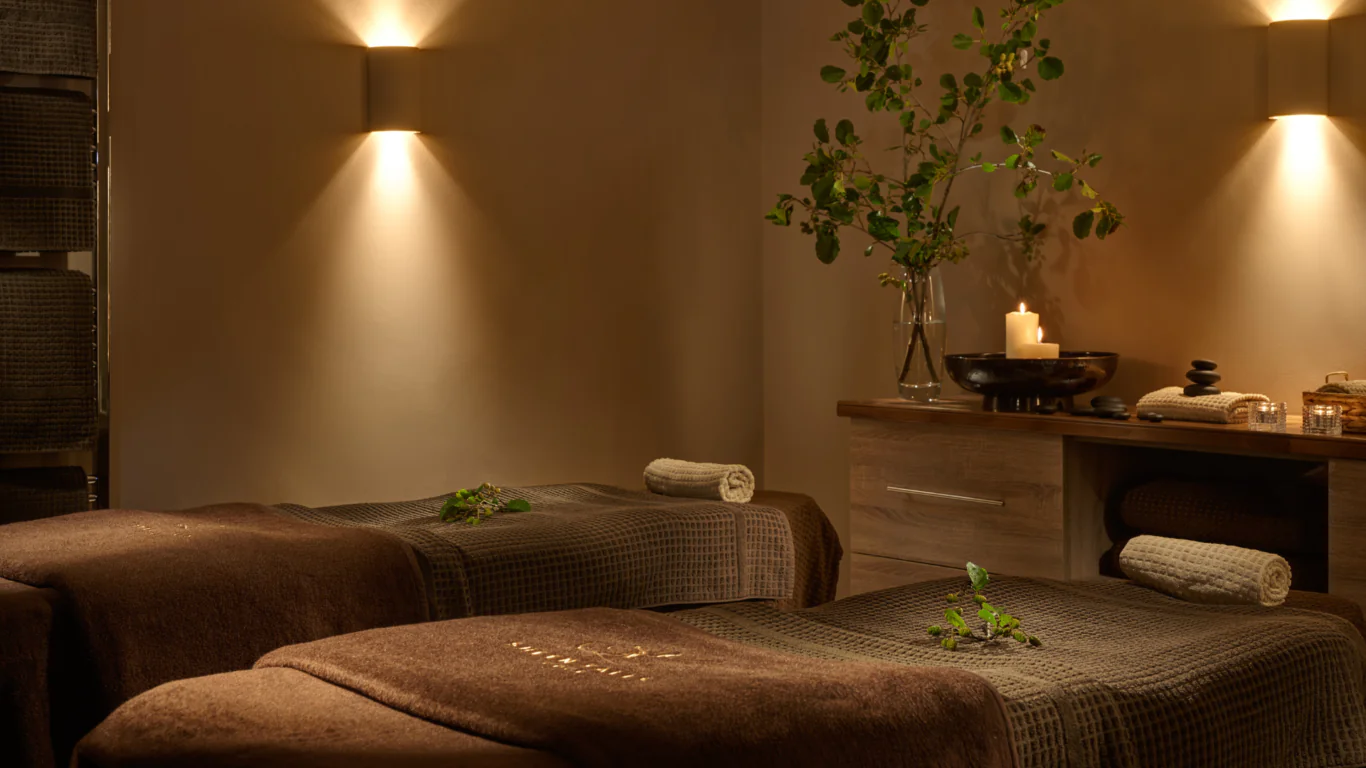 Tranquil spa room with candle-lit ambience, twin massage tables, soft brown linens, and relaxing greenery; perfect for relaxation.