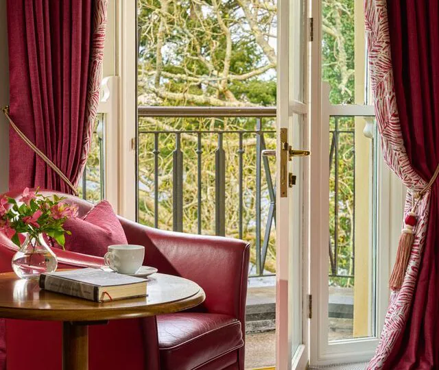 Cosy hotel room with red armchair, open balcony doors, scenic garden view, perfect for relaxing with a book and tea.