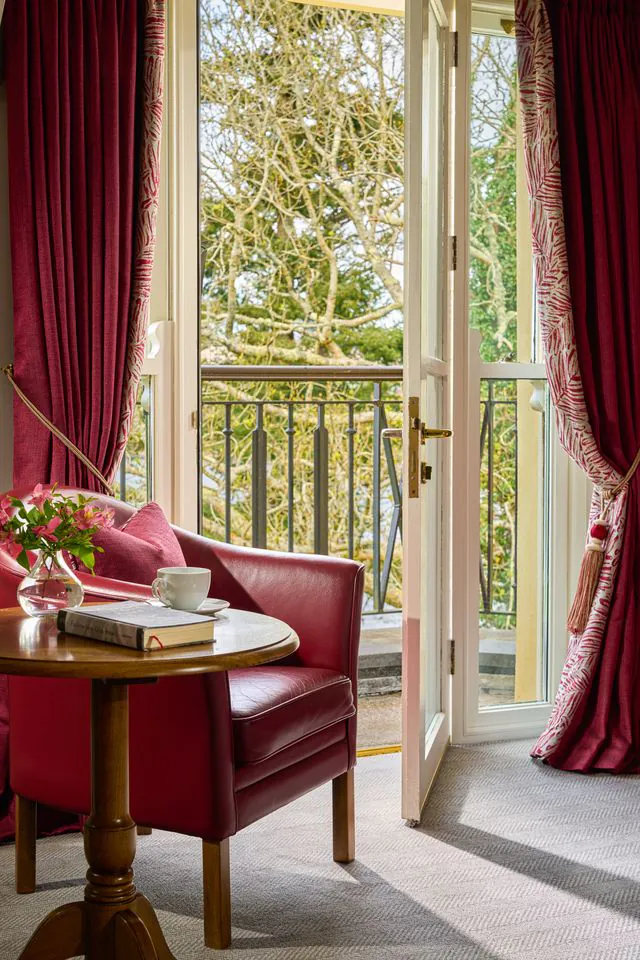 Cosy hotel room with red armchair, open balcony doors, scenic garden view, perfect for relaxing with a book and tea.