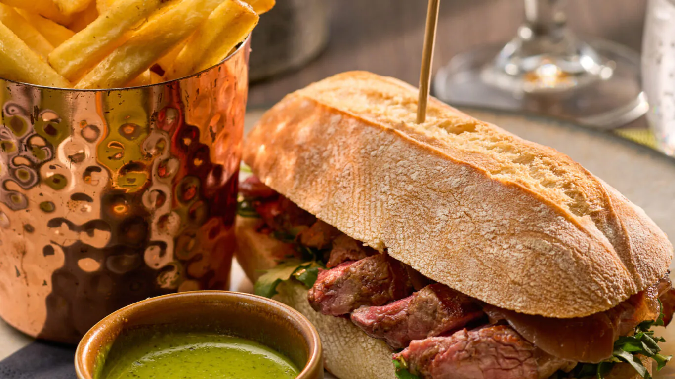 Enjoy gourmet steak sandwich with crispy chips and refreshing drink at a stylish hotel restaurant, ideal for relaxing dining.