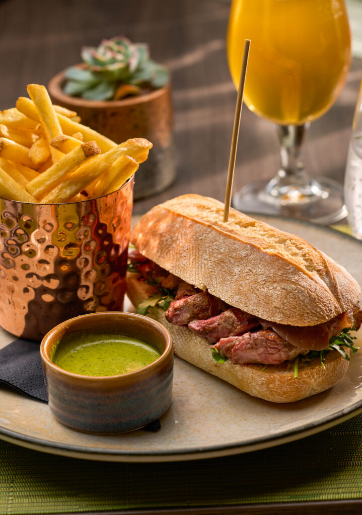 Savour a gourmet steak sandwich with crispy chips, green sauce, and a refreshing drink at our elegant hotel restaurant.