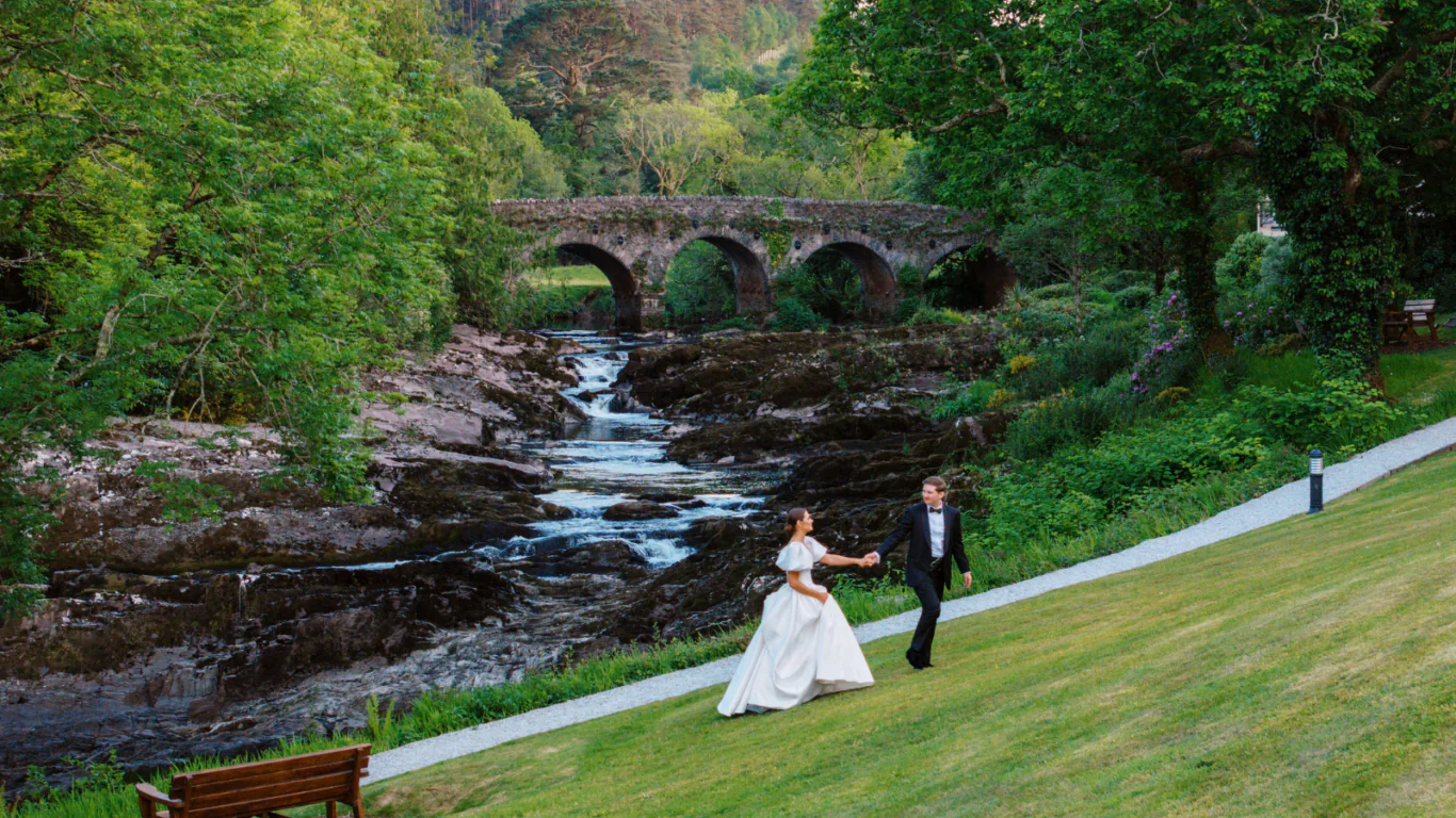 Romantic couple strolls by a picturesque stone bridge and serene river in lush green hotel garden. Perfect for weddings.