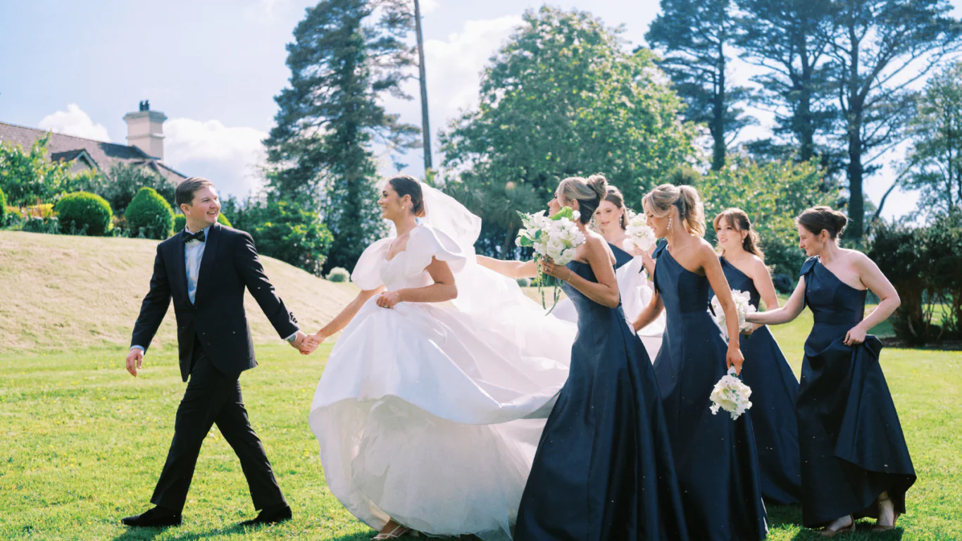 Bride and groom walk joyfully on lush hotel lawn with bridesmaids, scenic garden view, perfect for romantic weddings.