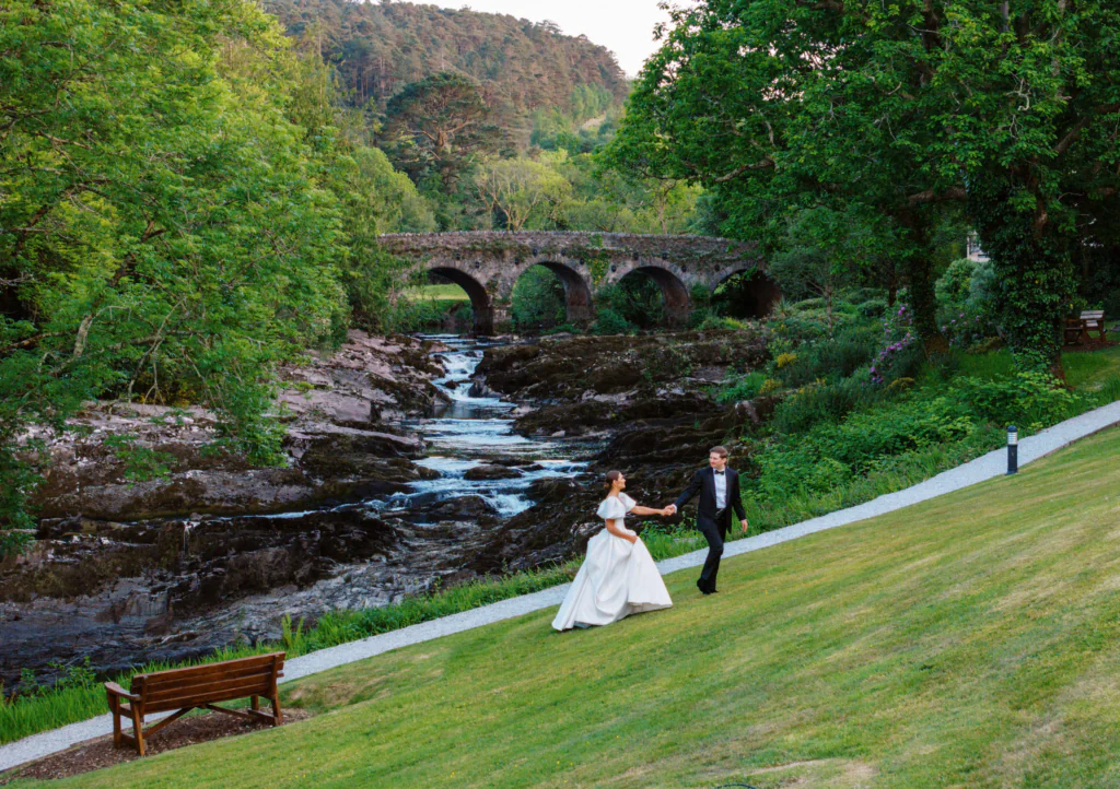Wedding couple walks on lush lawn by a stone bridge and tranquil river, surrounded by vibrant greenery.