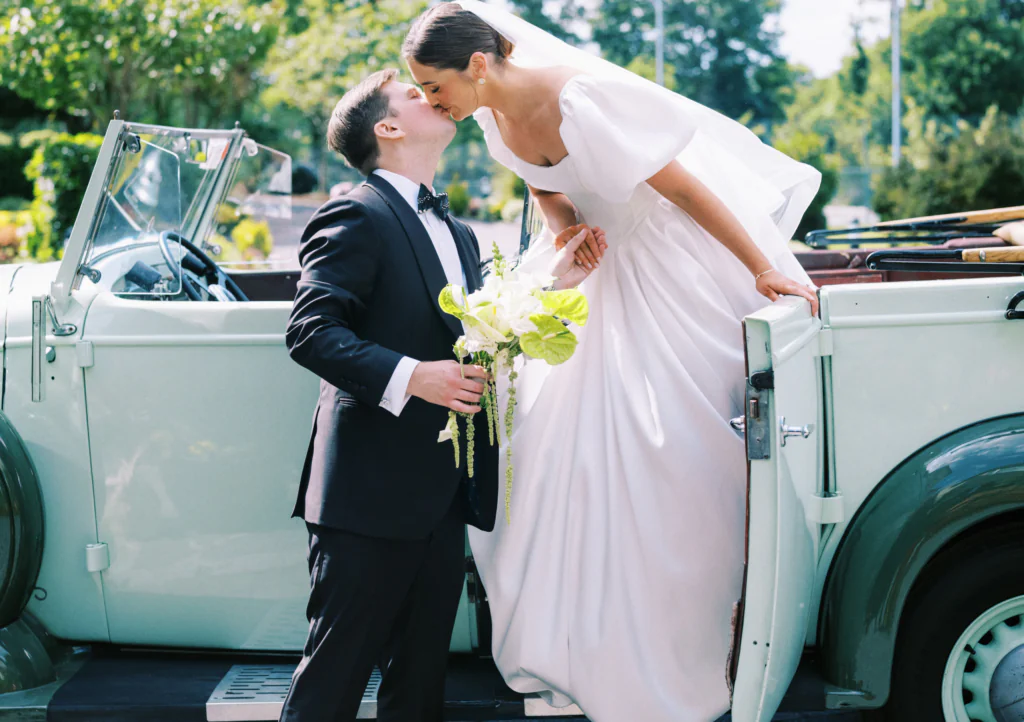 Newlyweds share a kiss by a classic car under sunny gardens, perfect for a romantic hotel wedding experience.