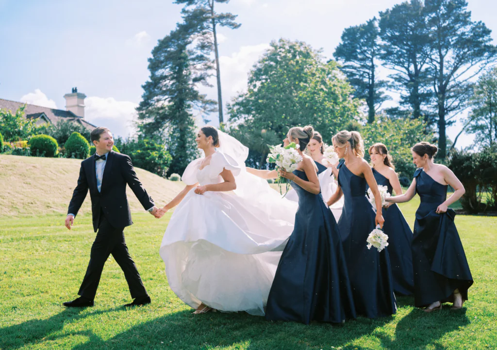 Bride in elegant gown and bridesmaids in navy dresses walking on lush lawn under clear sky at charming country hotel.