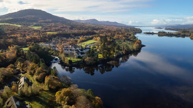 Scenic aerial view of idyllic lakeside hotel surrounded by lush autumn foliage and mountains, offering serene luxury and comfort.