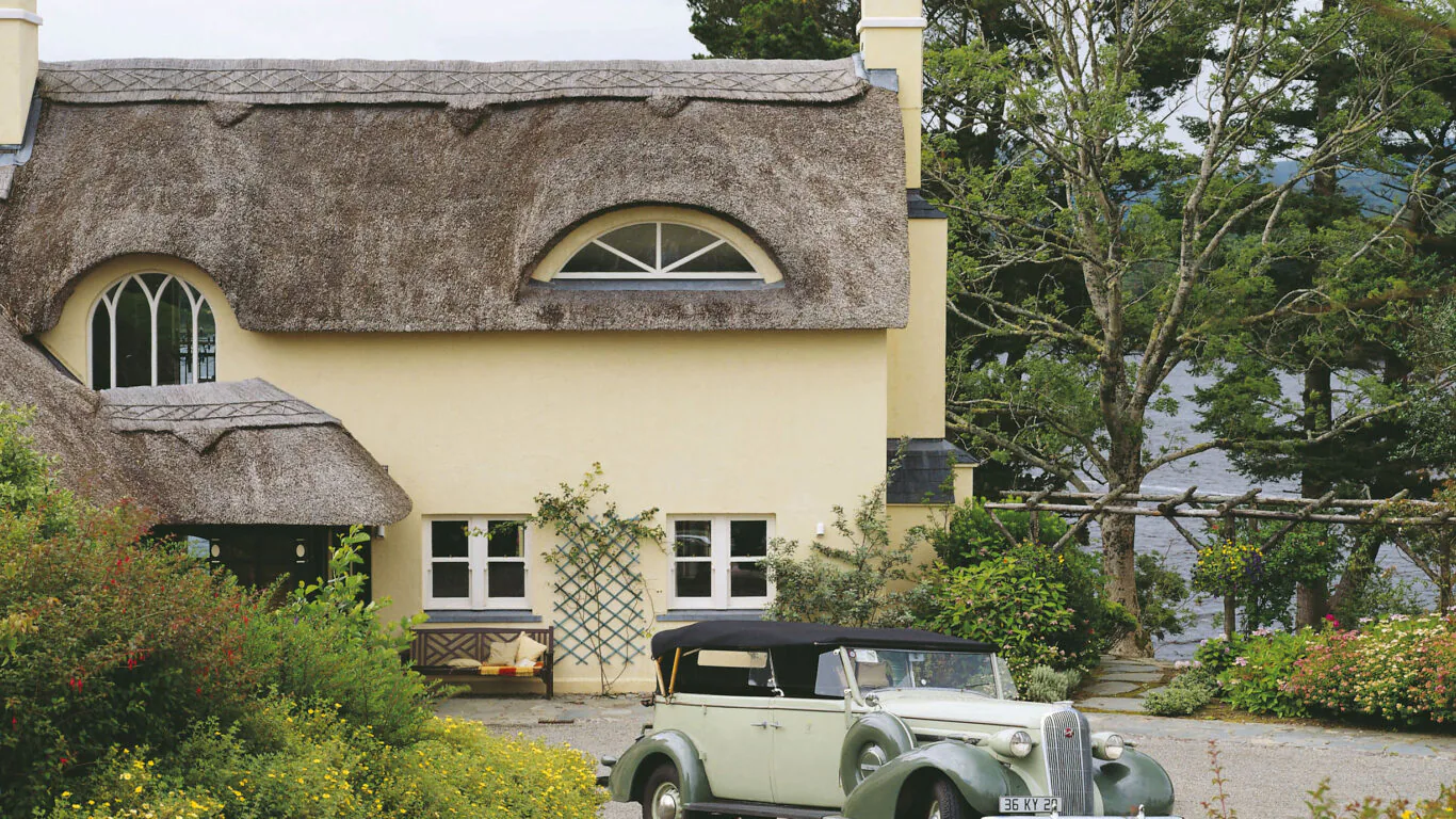 Charming thatched cottage with vintage car, lush gardens, and scenic lake view, offering a tranquil hotel retreat.
