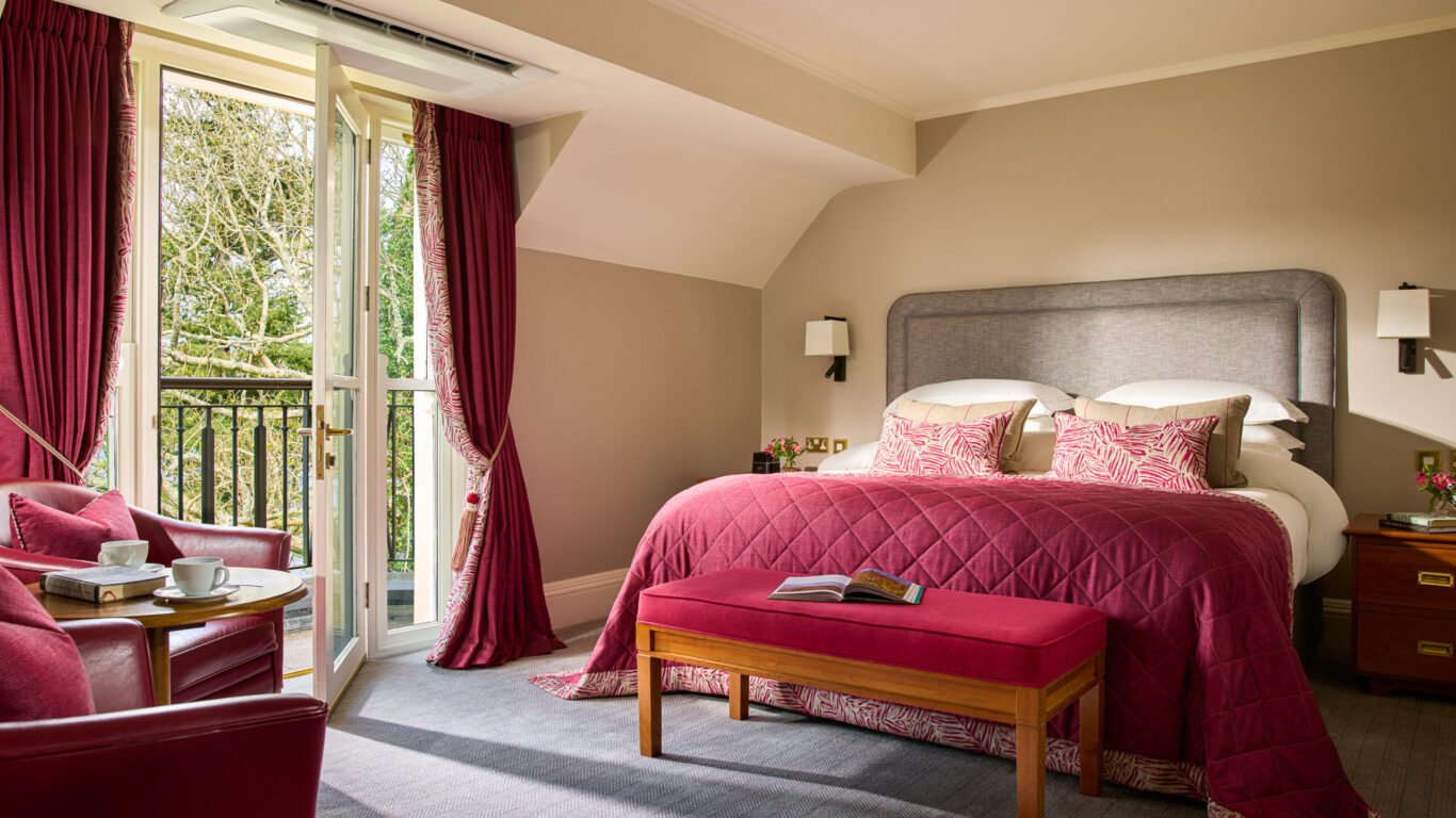 Elegant hotel room with plush king bed, balcony view, cosy seating, and luxurious red accents. Perfect for a relaxing stay.