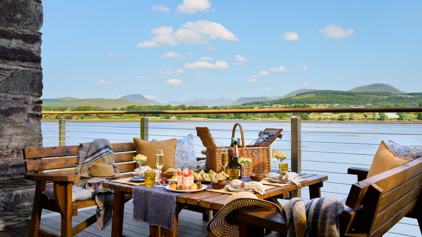 Scenic outdoor dining with a lakeside view, wooden table set with gourmet picnic, surrounded by mountains and clear skies.