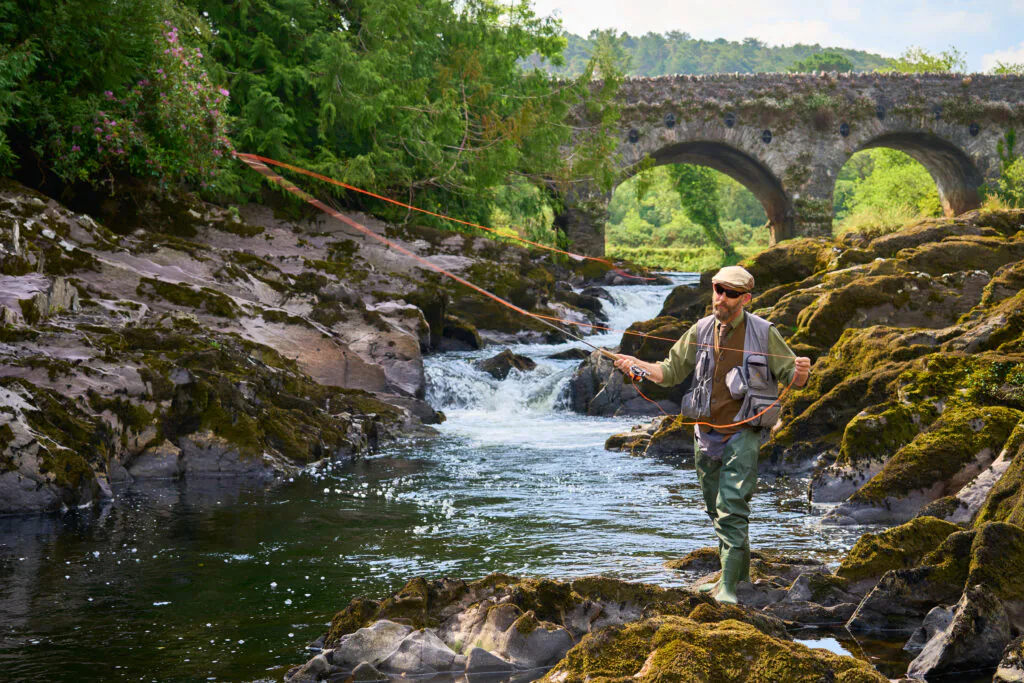 Guest enjoying fly fishing in scenic river with stone bridge and lush greenery, perfect for adventure and relaxation.