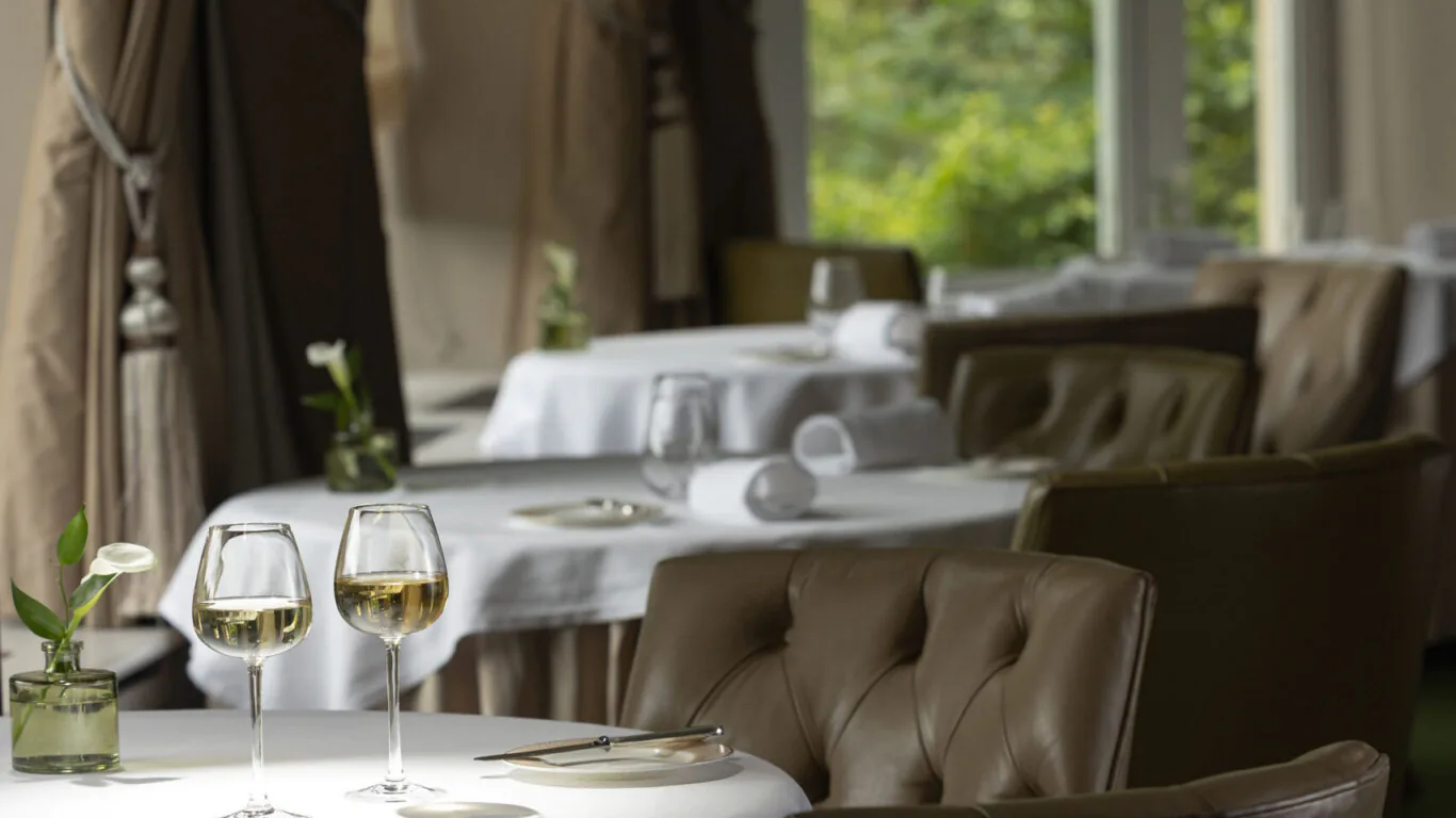 Elegant dining area with garden views, featuring white tablecloths, plush chairs, and glasses of wine for a tranquil meal.
