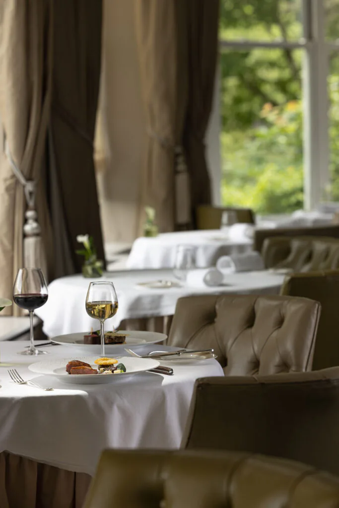 Elegant hotel dining room with wine, gourmet dishes, and garden view through large windows, offering a refined dining experience.