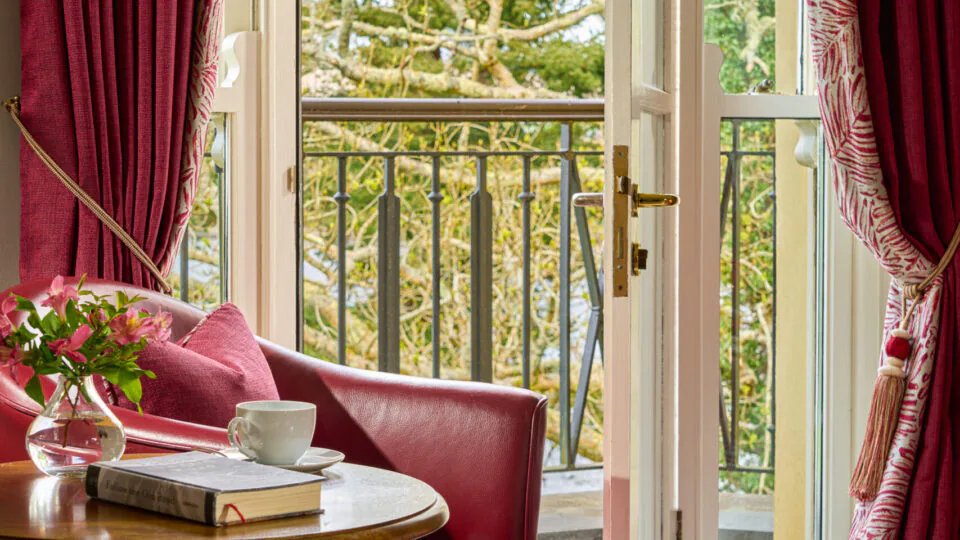 Cosy hotel room with red armchair, table, open balcony doors, and garden view, promising serene relaxation.